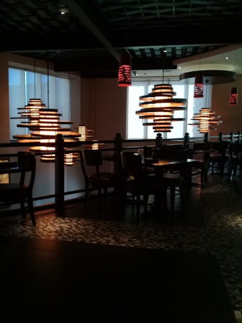 A dimly lit interior space featuring modern pendant lights with a layered design that emit a warm glow. The setting includes several tables and chairs arranged under the lights, with mosaic-style flooring visible. The ceiling has an architectural pattern.