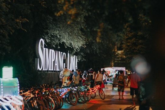 A lively nighttime scene with a crowd of people walking along a path lined with colorful illuminated bicycles. A large sign displaying 'Simpang Lima' is prominently featured, surrounded by lush greenery. The atmosphere is bustling and festive, with people interacting and enjoying the vibrant lights.