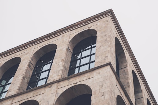 A building corner with large arched windows and a facade made of light-colored stone. The architecture features linear and geometric styling, giving a modern and minimalist appearance.