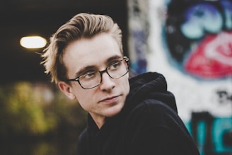 A young person wearing glasses and a black hoodie gazes pensively to the side, with a backdrop of blurred graffiti and light in the background.