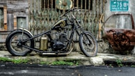 A custom motorcycle with a black and gold color scheme is parked in front of an old, rundown building with rusty metal gates. An old, rusted car door with graffiti is leaning against the wall to the right. There are green plants growing along the edge of the pavement, and a wet road is visible in the foreground.