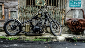 A custom motorcycle with a black and gold color scheme is parked in front of an old, rundown building with rusty metal gates. An old, rusted car door with graffiti is leaning against the wall to the right. There are green plants growing along the edge of the pavement, and a wet road is visible in the foreground.