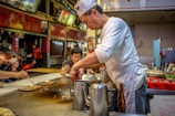 A street food vendor, wearing a white shirt and hat, prepares food on a hot surface while customers enjoy their meals. The setting is a bustling food stall with various cooking utensils and ingredients visible. Customers are seated nearby, some using chopsticks to eat their dishes.