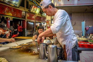 A street food vendor, wearing a white shirt and hat, prepares food on a hot surface while customers enjoy their meals. The setting is a bustling food stall with various cooking utensils and ingredients visible. Customers are seated nearby, some using chopsticks to eat their dishes.