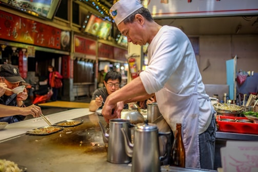 A street food vendor, wearing a white shirt and hat, prepares food on a hot surface while customers enjoy their meals. The setting is a bustling food stall with various cooking utensils and ingredients visible. Customers are seated nearby, some using chopsticks to eat their dishes.