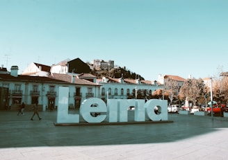 A large white sculpture of the word 'Leiria' is placed in an open plaza. People are casually walking around, and several cars are parked along the sides. In the background, there are traditional buildings with tiled roofs, and a hilltop castle can be seen under a clear blue sky.