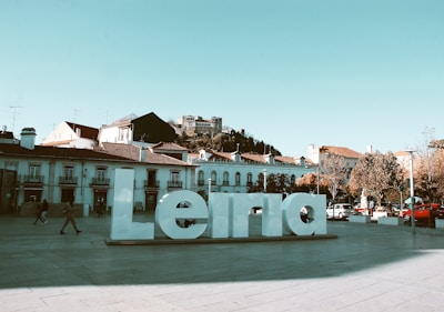 A large white sculpture of the word 'Leiria' is placed in an open plaza. People are casually walking around, and several cars are parked along the sides. In the background, there are traditional buildings with tiled roofs, and a hilltop castle can be seen under a clear blue sky.