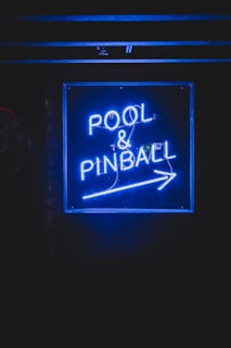 A bright blue neon sign is displayed in dark surroundings, featuring the words 'POOL & PINBALL' with an arrow pointing to the right.