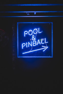 A bright blue neon sign is displayed in dark surroundings, featuring the words 'POOL & PINBALL' with an arrow pointing to the right.