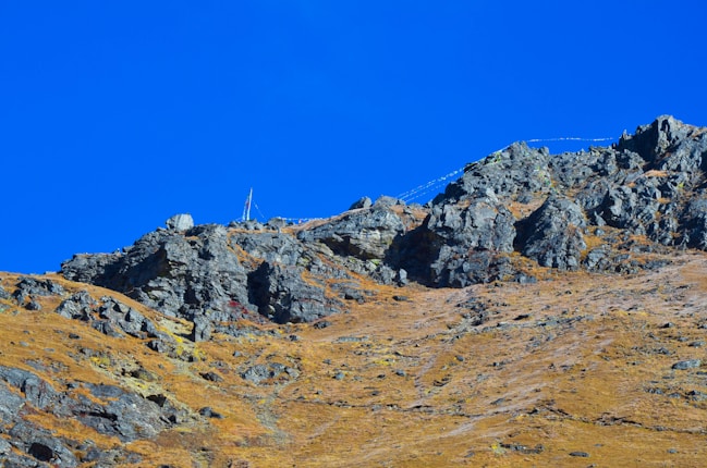 A rugged, rocky mountain landscape with a clear blue sky. The terrain is steep and scattered with large rocks, and there are prayer flags stretching across the top of the ridge.