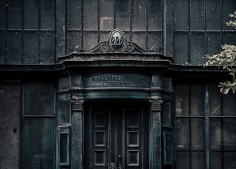 An old, weathered building facade with ornate architectural details. The words 'National Trust Company Limited' are inscribed above a large, dark wooden door. The structure appears abandoned, with dirty, grimy glass windows and a neglected appearance. Sparse foliage is visible to the side, adding to the decayed atmosphere.