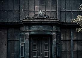 An old, weathered building facade with ornate architectural details. The words 'National Trust Company Limited' are inscribed above a large, dark wooden door. The structure appears abandoned, with dirty, grimy glass windows and a neglected appearance. Sparse foliage is visible to the side, adding to the decayed atmosphere.