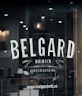 A barber shop window features the name 'BELGARD' prominently in large, bold letters. Below it, the words 'Barbier' and 'Barbershop & Spa' are displayed with decorative elements like straight razors. The reflection shows a dimly lit interior with a classic barber pole, a hanging vintage light bulb, and grooming products on a wooden shelf. The website www.belgardmtl.ca is also visible.