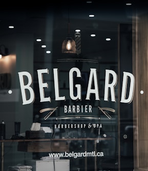 A barber shop window features the name 'BELGARD' prominently in large, bold letters. Below it, the words 'Barbier' and 'Barbershop & Spa' are displayed with decorative elements like straight razors. The reflection shows a dimly lit interior with a classic barber pole, a hanging vintage light bulb, and grooming products on a wooden shelf. The website www.belgardmtl.ca is also visible.