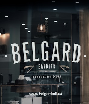 A barber shop window features the name 'BELGARD' prominently in large, bold letters. Below it, the words 'Barbier' and 'Barbershop & Spa' are displayed with decorative elements like straight razors. The reflection shows a dimly lit interior with a classic barber pole, a hanging vintage light bulb, and grooming products on a wooden shelf. The website www.belgardmtl.ca is also visible.