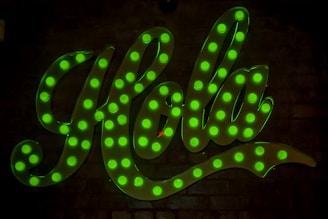 Large, cursive script spelling out 'Hola' with bright green, illuminated light bulbs on a dark background.