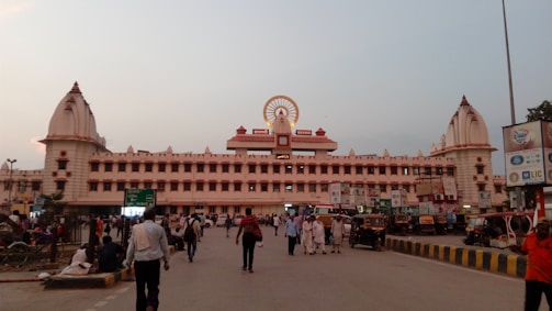A large building with distinct architectural features is visible, with a prominent clock and emblem at its center. The structure is flanked by two ornate tower-like elements on each side. Several people are walking in the foreground, while auto-rickshaws and other vehicles are parked near the building.