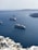A scenic view of the ocean featuring multiple cruise ships sailing. In the foreground, there are white buildings with blue accents located on a hillside. The background shows calm, blue waters extending towards distant, rocky landforms.
