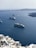 A scenic view of the ocean featuring multiple cruise ships sailing. In the foreground, there are white buildings with blue accents located on a hillside. The background shows calm, blue waters extending towards distant, rocky landforms.