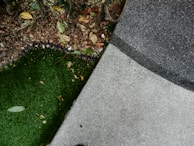 A scene consisting of green artificial grass on the left, dry leaves scattered around, and a textured gray concrete surface on the right. The area near the grass appears to have soil and is partially bordered by a black edge.