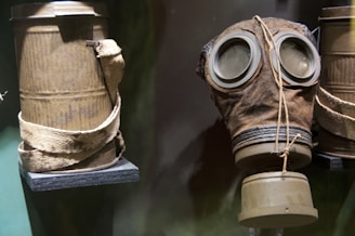 A vintage gas mask is displayed next to a cylindrical container with a worn strap. The materials appear aged with a gritty texture, suggesting historical use or an antique collection.