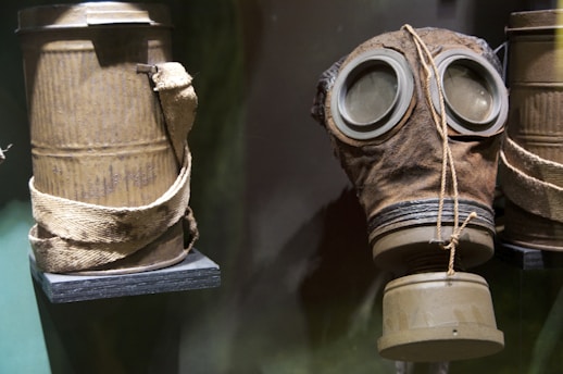 A vintage gas mask is displayed next to a cylindrical container with a worn strap. The materials appear aged with a gritty texture, suggesting historical use or an antique collection.