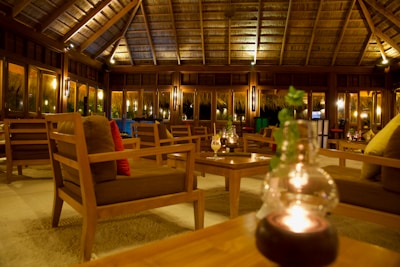 A cozy lounge area with wooden furniture sits under a thatched roof. Soft lighting creates a warm and inviting atmosphere, with several tables and cushioned chairs arranged neatly. Candle-lit glass decorations and green plants add to the relaxed ambiance.