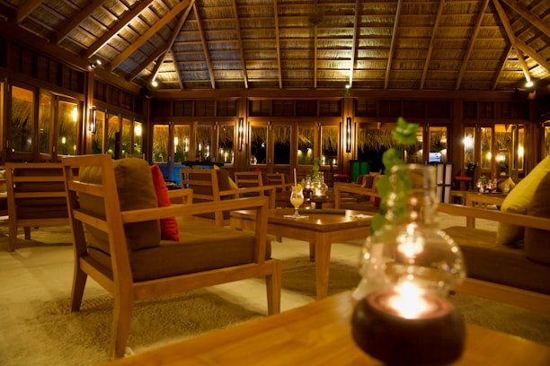 A cozy lounge area with wooden furniture sits under a thatched roof. Soft lighting creates a warm and inviting atmosphere, with several tables and cushioned chairs arranged neatly. Candle-lit glass decorations and green plants add to the relaxed ambiance.