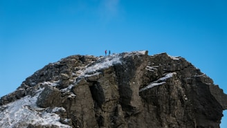 A rocky mountain peak with patches of snow is depicted under a clear blue sky. Two people are standing at the summit, indicating a sense of achievement and adventure.