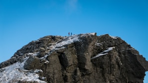 A rocky mountain peak with patches of snow is depicted under a clear blue sky. Two people are standing at the summit, indicating a sense of achievement and adventure.