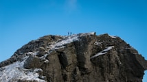 A rocky mountain peak with patches of snow is depicted under a clear blue sky. Two people are standing at the summit, indicating a sense of achievement and adventure.
