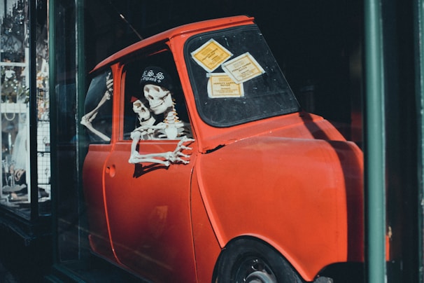 A display featuring a red car with skeleton figures inside, positioned as driver and passenger, with a quirky and humorous intent. The car is situated in a glass storefront, and there are notices attached to the window.
