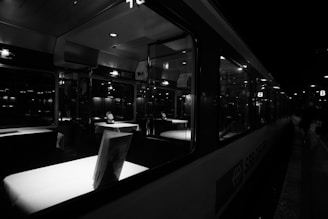 An interior view of a train dining car illuminated by soft overhead lights. The tables are covered with white tablecloths, accented by decorative settings. The windows reflect the dim lighting from outside, and the car is mostly empty, suggesting a calm, quiet atmosphere.