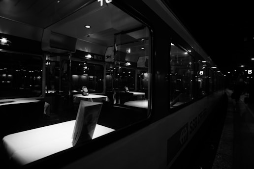 An interior view of a train dining car illuminated by soft overhead lights. The tables are covered with white tablecloths, accented by decorative settings. The windows reflect the dim lighting from outside, and the car is mostly empty, suggesting a calm, quiet atmosphere.