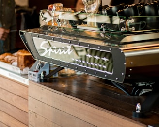 A modern espresso machine branded 'Spirit' with the model name 'Triplette' is prominently displayed on a wooden countertop. Several black mugs are placed on top of the machine. In the background, there are some bread loaves and packaged snacks like chia bars.