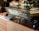 A modern espresso machine branded 'Spirit' with the model name 'Triplette' is prominently displayed on a wooden countertop. Several black mugs are placed on top of the machine. In the background, there are some bread loaves and packaged snacks like chia bars.