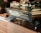 A modern espresso machine branded 'Spirit' with the model name 'Triplette' is prominently displayed on a wooden countertop. Several black mugs are placed on top of the machine. In the background, there are some bread loaves and packaged snacks like chia bars.