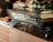 A modern espresso machine branded 'Spirit' with the model name 'Triplette' is prominently displayed on a wooden countertop. Several black mugs are placed on top of the machine. In the background, there are some bread loaves and packaged snacks like chia bars.
