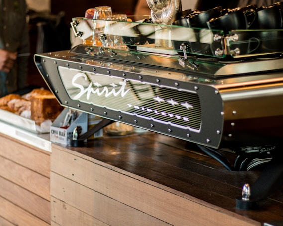 A modern espresso machine branded 'Spirit' with the model name 'Triplette' is prominently displayed on a wooden countertop. Several black mugs are placed on top of the machine. In the background, there are some bread loaves and packaged snacks like chia bars.