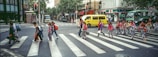 A group of school children wearing uniforms and backpacks cross a wide street at a pedestrian crosswalk in an urban area. The street is lined with buildings, including a convenience store and various shops. Several vehicles, including a yellow minibus and a green bus, are visible in the background. Traffic lights indicate it's a safe time to cross, and there is some greenery along the street.