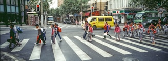 A group of school children wearing uniforms and backpacks cross a wide street at a pedestrian crosswalk in an urban area. The street is lined with buildings, including a convenience store and various shops. Several vehicles, including a yellow minibus and a green bus, are visible in the background. Traffic lights indicate it's a safe time to cross, and there is some greenery along the street.