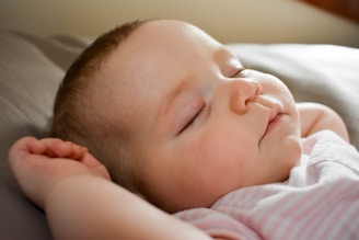 A peaceful sleeping baby with soft, closed eyes and a gentle expression, lying on a bed. The baby's head is slightly tilted, and one arm is raised above the head. The lighting is soft, highlighting the smoothness of the baby's skin and creating a serene atmosphere.