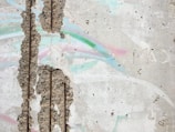 A textured concrete wall with large patches of exposed aggregate. There are pastel-colored streaks of pink, green, and blue across the surface. The wall has several vertical indents and appears weathered. Some graffiti markings, including a small heart, can be seen on the wall.