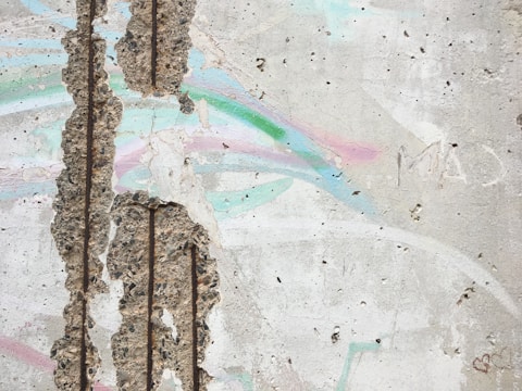 A textured concrete wall with large patches of exposed aggregate. There are pastel-colored streaks of pink, green, and blue across the surface. The wall has several vertical indents and appears weathered. Some graffiti markings, including a small heart, can be seen on the wall.