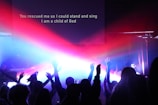 A group of people with raised hands in a dimly lit room, illuminated by vibrant beams of red, blue, and purple lights. A projection screen displays the text 'You rescued me so I could stand and sing I am a child of God.'