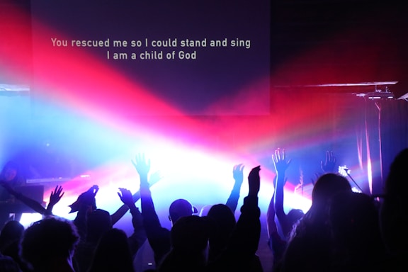 A group of people with raised hands in a dimly lit room, illuminated by vibrant beams of red, blue, and purple lights. A projection screen displays the text 'You rescued me so I could stand and sing I am a child of God.'