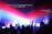 A group of people with raised hands in a dimly lit room, illuminated by vibrant beams of red, blue, and purple lights. A projection screen displays the text 'You rescued me so I could stand and sing I am a child of God.'