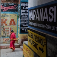 A narrow street in Varanasi filled with colorful signage on the walls advertising cafes, restaurants, yoga, and music schools. A woman in a bright pink saree walks through the lane.