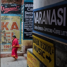 A narrow street in Varanasi filled with colorful signage on the walls advertising cafes, restaurants, yoga, and music schools. A woman in a bright pink saree walks through the lane.
