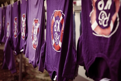 A line of purple shirts are hanging, each featuring a circular logo with the letters 'R' and 'P' surrounded by a red and white design. The shirts appear to be drying outside.